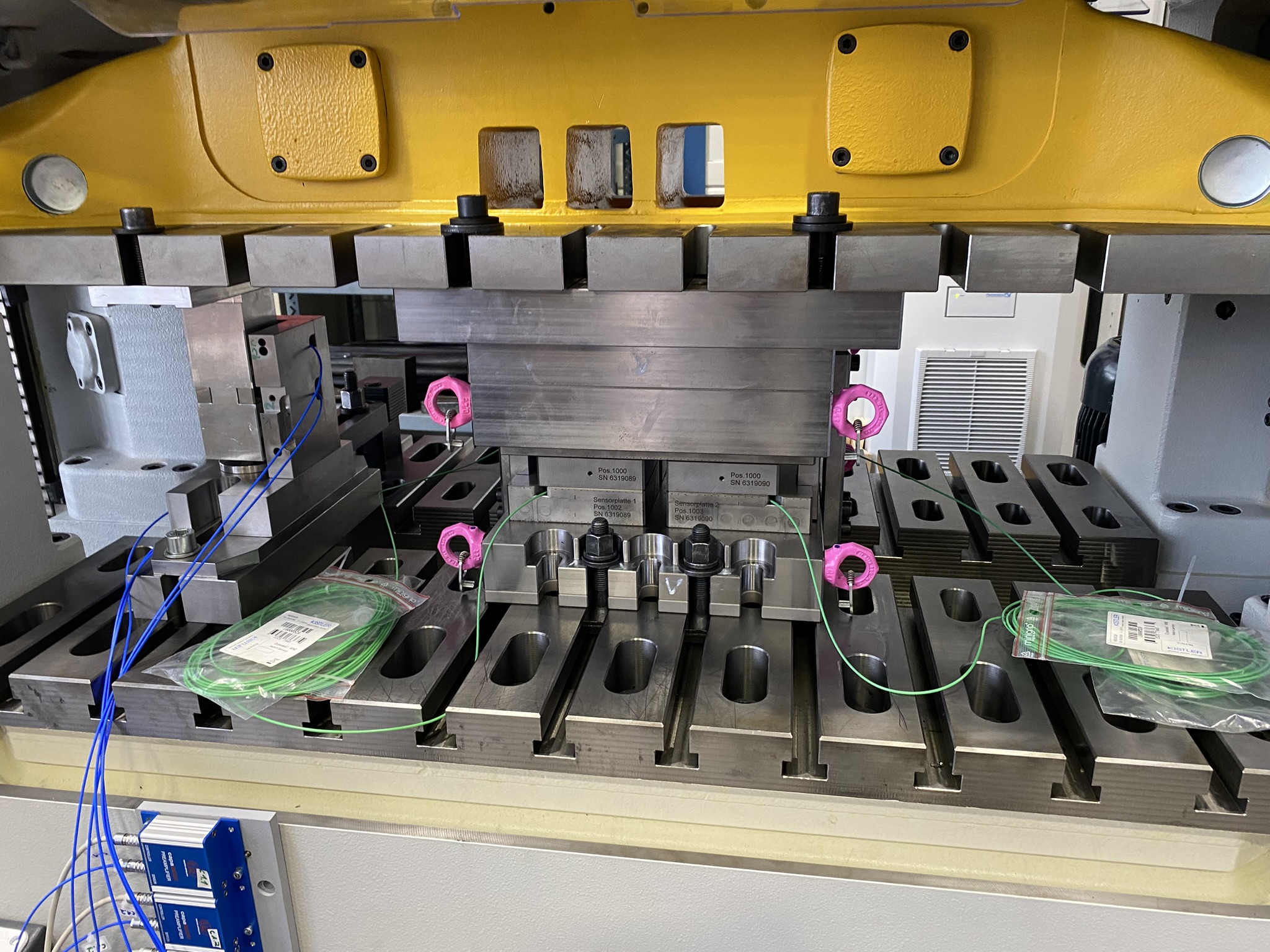 Einbau des entwickelten Systems zur dynamischen Charakterisierung von Schnellläuferpressen. Links im Presseneinbauraum: Messmodul mit fünf kapazitiven Sensoren (blaue Kabel) zur Überwachung des Presseneinbauraums während der Prüfroutine. Zentral im Presseneinbauraum: Werkzeugmodul zur Lasteinleitung mit vier piezoelektrischen Kraftmessdosen (grüne Kabel) zur Überwachung der Kraftrandbedingungen während der Prüfroutine. (Quelle: Technische Universität München, Lehrstuhl für Umformtechnik und Gießereiwesen (utg))